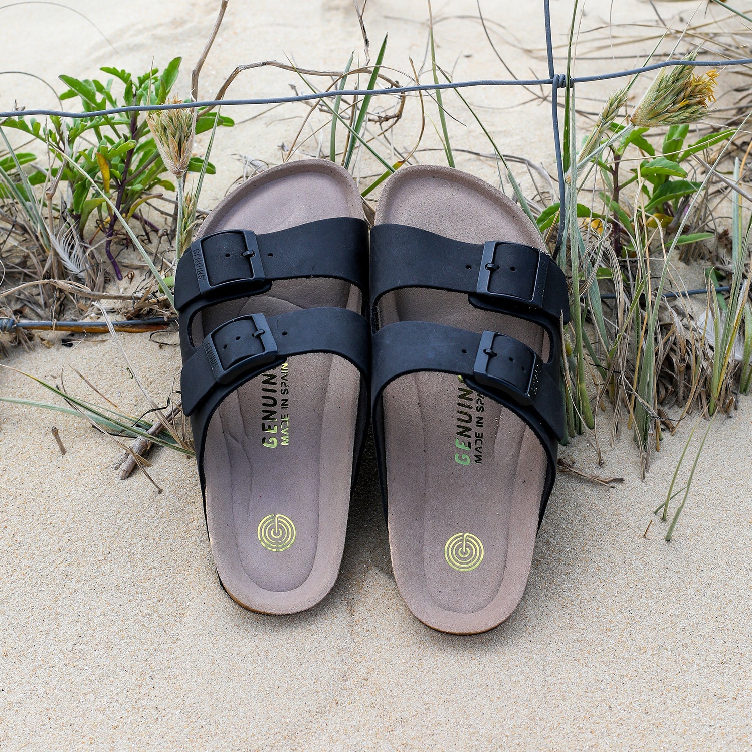 Black leather sandal with double foot strap and faux fur foot bed leaning against a fence at the beach